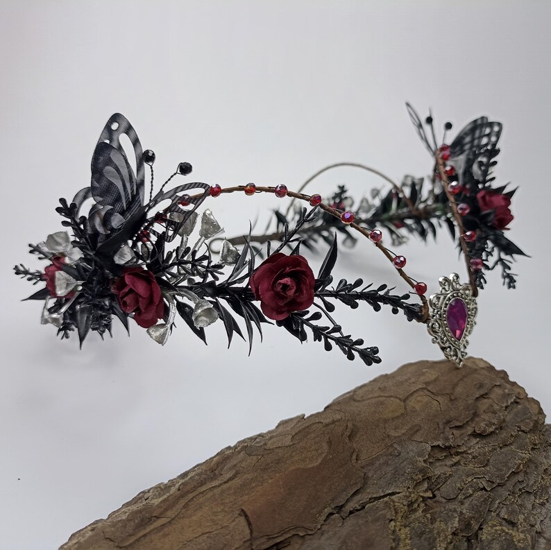 Pu&ograve; includere: Una corona floreale nera e argentata con rose rosse e un accento a farfalla. La corona &egrave; adornata con piccole perle d'argento e una gemma rosa.