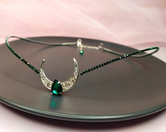 May include: A silver crescent moon headband with a green gemstone in the center. The headband is made of small green beads.