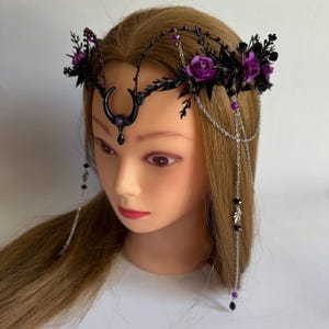 May include: A headpiece featuring a black crescent moon centerpiece with a purple gem, accented by dark foliage, purple roses, and silver chains. The design includes dangling silver chains with black and purple beads that drape along the sides.