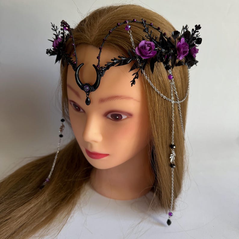 May include: A headpiece featuring a black crescent moon centerpiece with a purple gem, accented by dark foliage, purple roses, and silver chains. The design includes dangling silver chains with black and purple beads that drape along the sides.