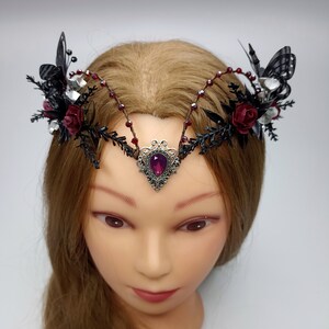 Pu&ograve; includere: Una corona nera e argentata con rose rosse e farfalle nere. La corona ha una pietra preziosa viola al centro.