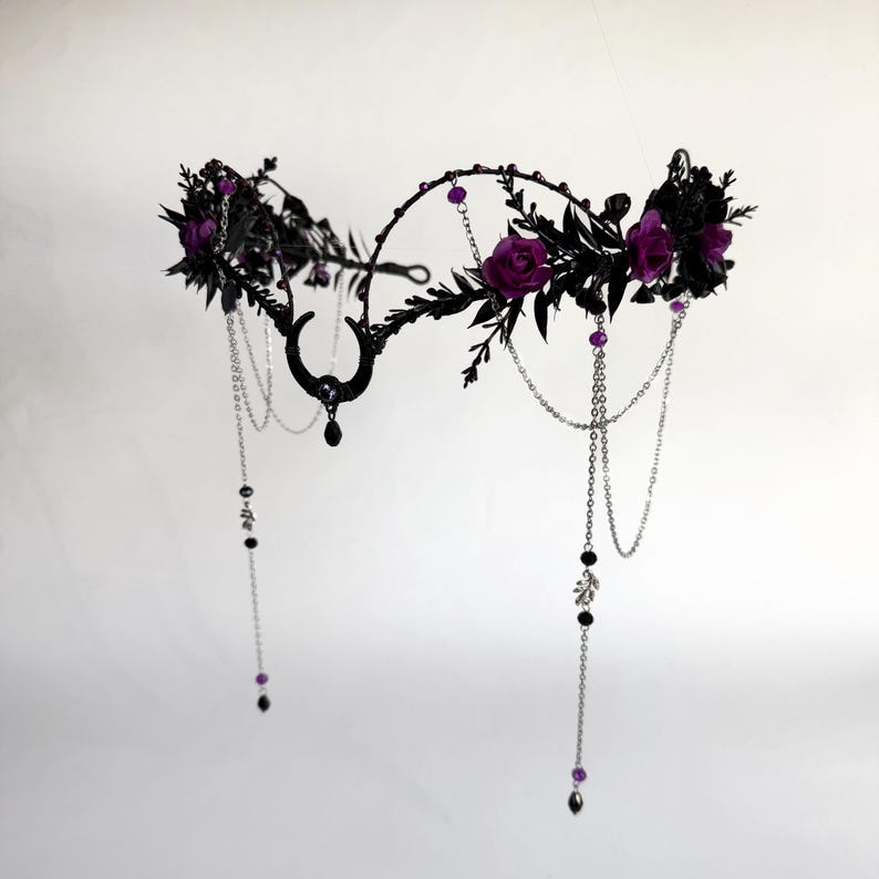 May include: A headpiece featuring a black metal frame adorned with dark leaves, deep purple roses, and a central crescent moon charm. The design includes delicate silver-toned chains draped with small black and purple beads and teardrop accents.