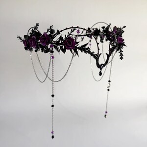 May include: A headpiece featuring dark purple roses and black foliage arranged on a circular frame. The design includes silver-toned chains draped downwards, accented with small black and purple beads, and a central black crescent moon charm.