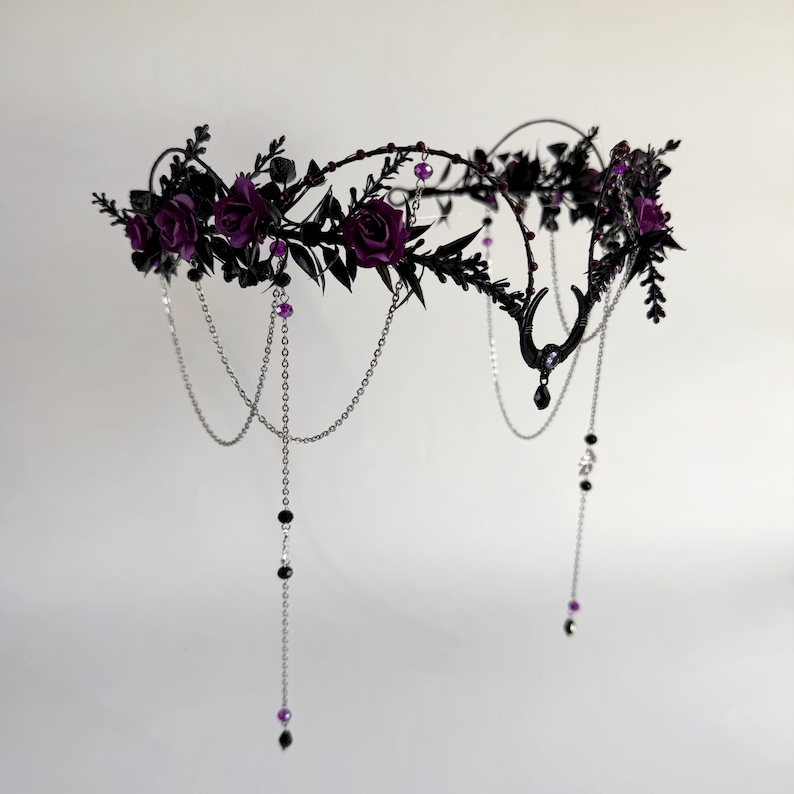 May include: A headpiece featuring dark purple roses and black foliage arranged on a circular frame. The design includes silver-toned chains draped downwards, accented with small black and purple beads, and a central black crescent moon charm.
