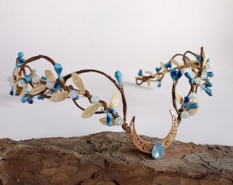 May include: A gold wire headband with blue and white beads and a crescent moon charm. The headband is decorated with gold leaves and blue berries.