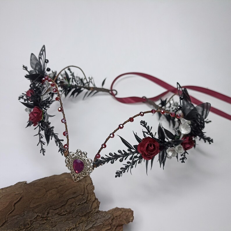 Pu&ograve; includere: Una corona floreale nera e bordeaux con un accento di gioiello. La corona &egrave; realizzata con fiori neri, rose bordeaux e farfalle nere. &Egrave; adornata con perline d'argento e un'unica grande gemma rosa.