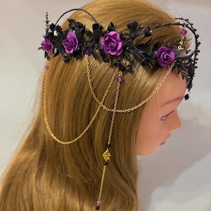 May include: A headpiece featuring purple roses and black foliage arranged on a dark band. The accessory includes gold-toned chains that drape down the sides, accented with small purple beads, black crystals, and a gold-colored leaf charm.