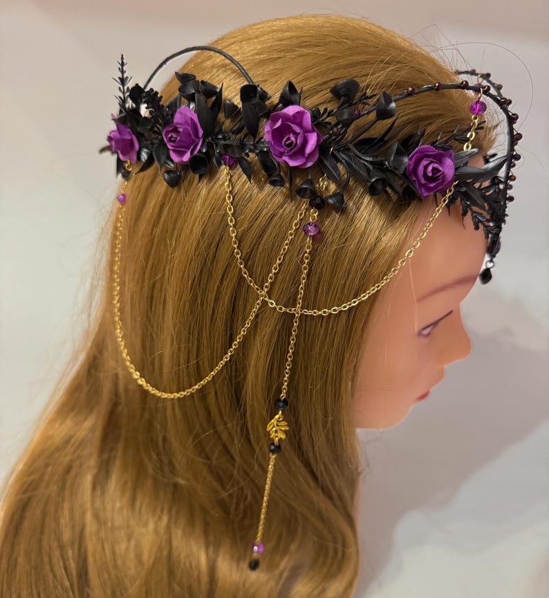 May include: A headpiece featuring purple roses and black foliage arranged on a dark band. The accessory includes gold-toned chains that drape down the sides, accented with small purple beads, black crystals, and a gold-colored leaf charm.