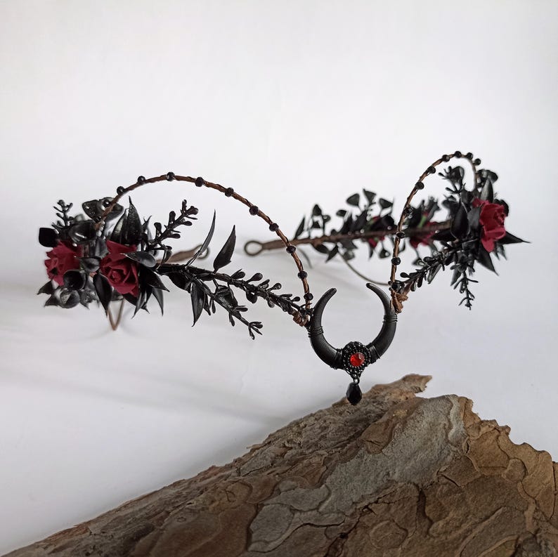 Peut inclure: Couronne florale noire avec des roses rouges et un charme de croissant de lune avec une pierre pr&eacute;cieuse rouge. La couronne est faite de fil de fer et est d&eacute;cor&eacute;e de feuilles et de baies noires.