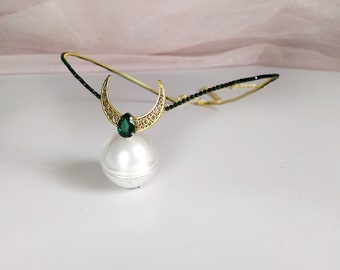 May include: A gold crescent moon headband with a green gemstone and a white pearl orb. The headband is made of black beads.