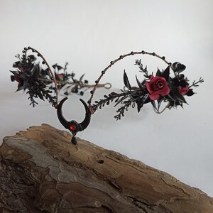 Peut inclure: Couronne florale noire avec des roses rouges et un charme de croissant de lune avec une pierre pr&eacute;cieuse rouge. La couronne est faite de fil de fer et est d&eacute;cor&eacute;e de feuilles et de baies noires.
