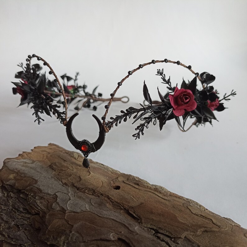 Peut inclure: Couronne florale noire avec des roses rouges et un charme de croissant de lune avec une pierre pr&eacute;cieuse rouge. La couronne est faite de fil de fer et est d&eacute;cor&eacute;e de feuilles et de baies noires.