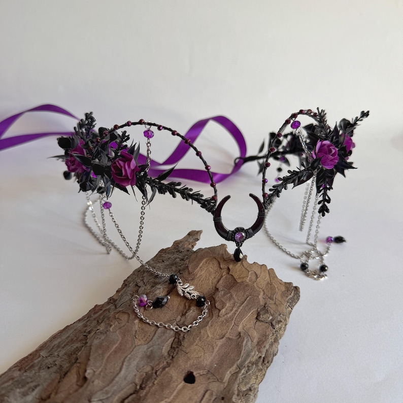 May include: A headpiece featuring a central black crescent moon charm with a purple stone, adorned with dark foliage, purple roses, silver chains, and small purple beads. The accessory includes long purple ribbons for tying and delicate metal accents.