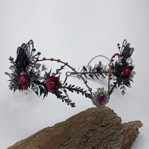 Pu&ograve; includere: Una corona floreale nera e argentata con rose rosse e un ciondolo argentato con una pietra preziosa rossa. La corona &egrave; realizzata in filo metallico ed &egrave; decorata con piume nere e perline argentate.