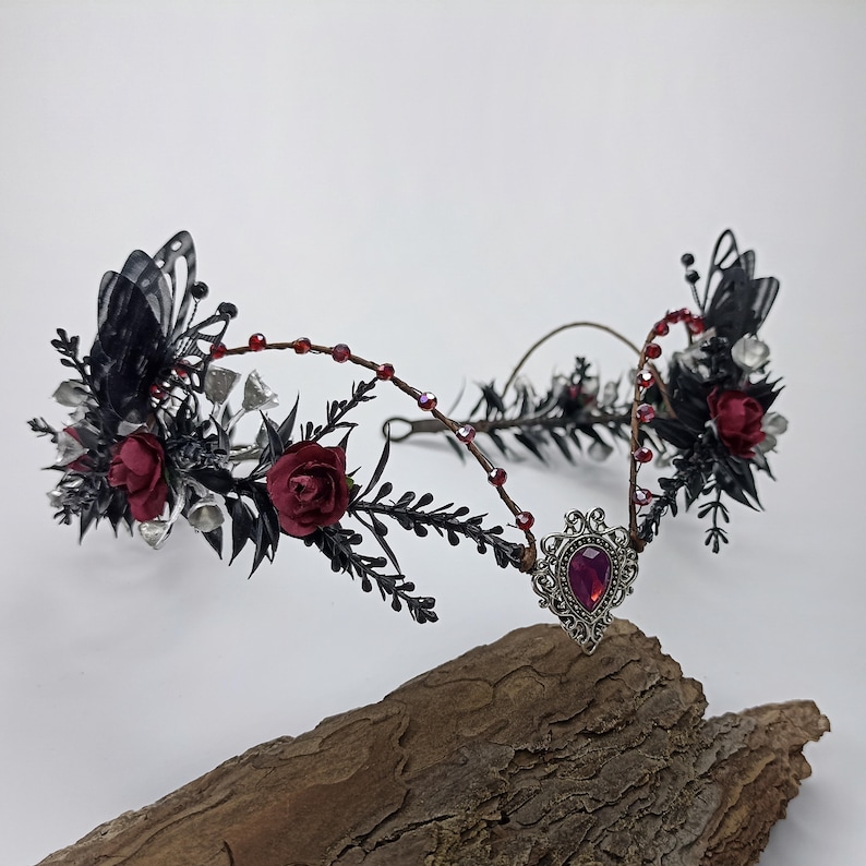 Pu&ograve; includere: Una corona floreale nera e argentata con rose rosse e un ciondolo argentato con una pietra preziosa rossa. La corona &egrave; realizzata in filo metallico ed &egrave; decorata con piume nere e perline argentate.