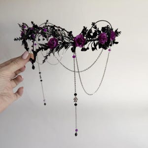 May include: A headpiece featuring a black wire frame adorned with dark foliage and deep purple roses. The accessory includes delicate silver-toned chains that drape downward, accented with small black beads and purple crystal-like spherical charms.