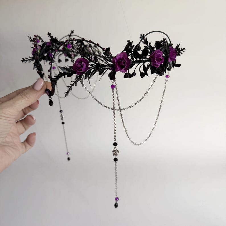 May include: A headpiece featuring a black wire frame adorned with dark foliage and deep purple roses. The accessory includes delicate silver-toned chains that drape downward, accented with small black beads and purple crystal-like spherical charms.