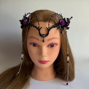 May include: A black headpiece featuring a central crescent moon charm with a purple gem and a dangling black bead. The design includes dark wire branches, purple fabric roses, and silver-toned chains with small leaf charms and purple beads.