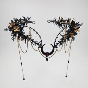 Puede incluir: Una corona floral negra y dorada con un creciente de luna y un acento de piedra preciosa roja. La corona está adornada con cadenas negras y doradas y cuentas negras.