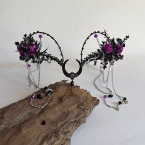May include: A black metal headpiece featuring a central crescent moon charm with a purple stone, accented by dark purple roses, black foliage, silver chains, and dangling purple and black beads. The headpiece has a symmetrical, arched design.