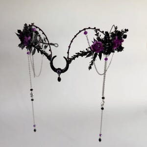 May include: A black metal headpiece featuring a central crescent moon charm with a small purple bead and a dangling black teardrop. The frame is adorned with dark leaves, deep purple roses, and thin silver chains with purple and black beads.