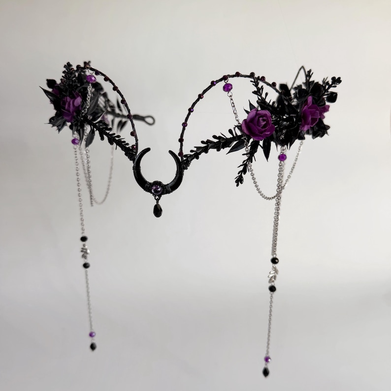 May include: A black metal headpiece featuring a central crescent moon charm with a small purple bead and a dangling black teardrop. The frame is adorned with dark leaves, deep purple roses, and thin silver chains with purple and black beads.