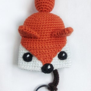 May include: A crocheted fox keychain with a white face, orange body, and brown tail. The keychain has a silver metal ring.