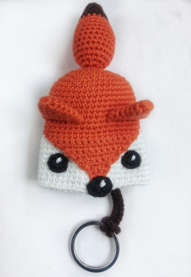 May include: A crocheted fox keychain with a white face, orange body, and brown tail. The keychain has a silver metal ring.