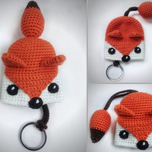 May include: A crocheted fox keychain with a white and orange body, black eyes, and a brown tail. The fox is attached to a silver key ring.