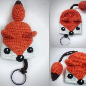 May include: A crocheted fox keychain with a white face, orange body, and brown tail. The fox has black eyes and a black nose.