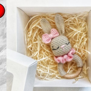 Puede incluir: Un sonajero de conejo de crochet con un lazo rosa y un anillo de madera. El sonajero está en una caja de regalo blanca con papel de seda.