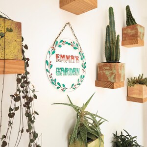 May include: A clear, oval-shaped sign with the text "EMMA'S GARDEN" in red and green, surrounded by a green leaf and berry design. The sign hangs from a chain. Various potted plants and wooden shelves are mounted on the white wall.