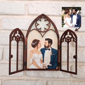 May include: A wooden frame with three arched windows, each with intricate carvings. The center window displays a painted portrait of a couple in wedding attire, while the side windows are open. The frame is mounted on a white brick wall.