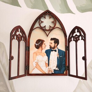 May include: A wedding portrait of a couple in a wooden frame with a gothic arch design. The frame is open and the couple is looking at each other. The woman is wearing a white wedding dress and the man is wearing a blue suit.