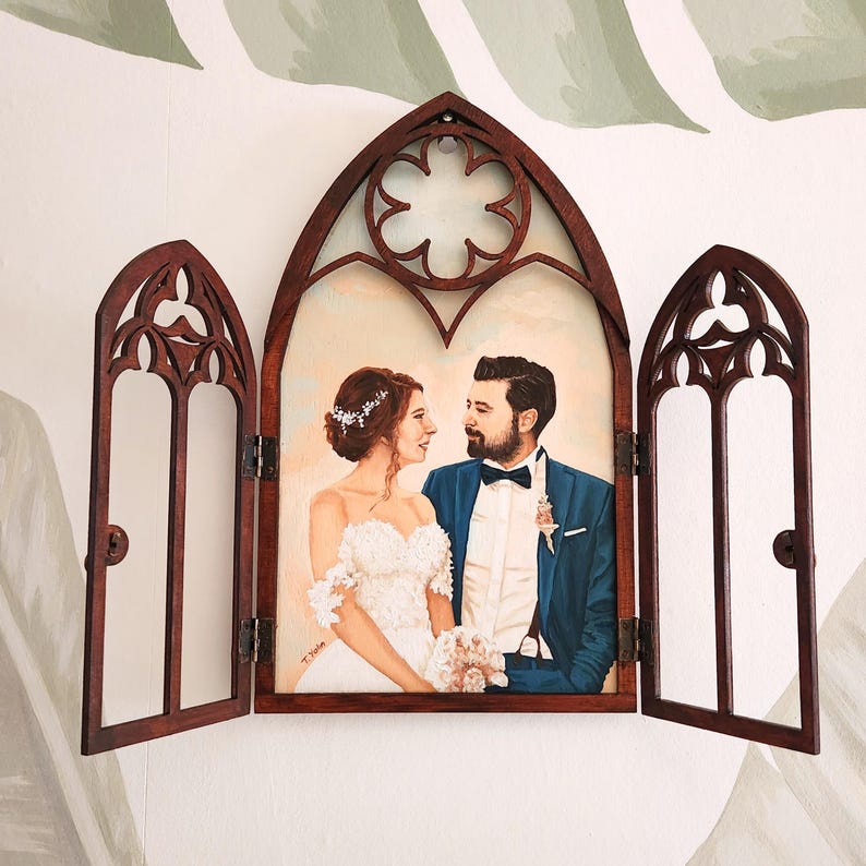 May include: A wedding portrait of a couple in a wooden frame with a gothic arch design. The frame is open and the couple is looking at each other. The woman is wearing a white wedding dress and the man is wearing a blue suit.