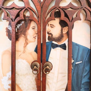 May include: A painted portrait of a couple in a wooden frame with a carved design. The woman is wearing a white dress and the man is wearing a blue suit with a white shirt and a black bow tie. They are looking at each other.