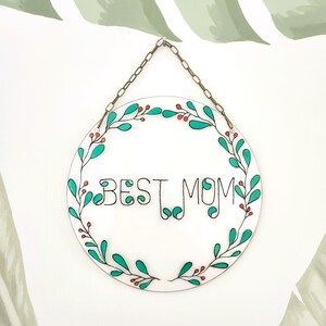 May include: A round, white sign with a gold chain for hanging. The sign has a green leafy wreath with red berries around the edge. The text "BEST MOM" is written in black inside the wreath.