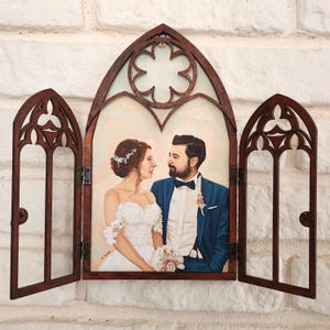 May include: A wedding portrait of a couple framed in a wooden archway with a decorative design. The couple is dressed in formal attire, the woman in a white gown and the man in a blue suit. The background is a soft, pastel sky.
