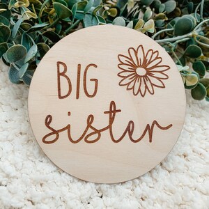 May include: A round wooden sign with the words "BIG sister" and a stylized daisy design.