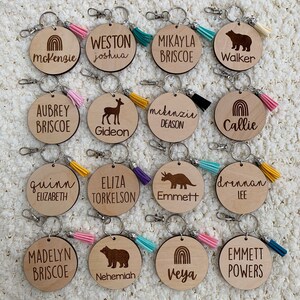 May include: Twelve wooden keychains with different designs and names engraved on them. Each keychain has a small tassel attached to it. The designs include a rainbow, a deer, a bear, a dinosaur, and a mountain range. The names include McKenzie, Weston, Mikayla, Walker, Aubrey, Gideon, McKenzie, Callie, Quinn, Eliza, Emmett, Drennan, Madelyn, Nehemiah, Veya, and Emmett.