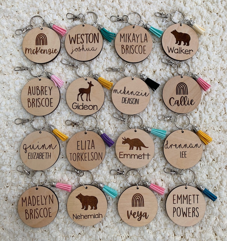 May include: Twelve wooden keychains with different designs and names engraved on them. Each keychain has a small tassel attached to it. The designs include a rainbow, a deer, a bear, a dinosaur, and a mountain range. The names include McKenzie, Weston, Mikayla, Walker, Aubrey, Gideon, McKenzie, Callie, Quinn, Eliza, Emmett, Drennan, Madelyn, Nehemiah, Veya, and Emmett.
