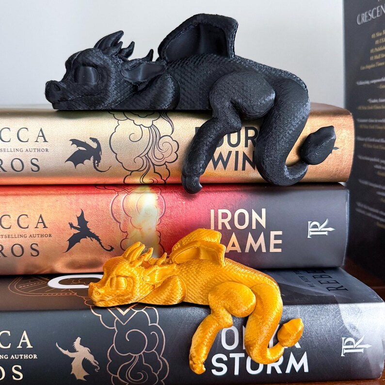 May include: Two 3D printed dragon bookends, one black and one gold, are resting on top of three books. The books are titled "A Storm of Swords", "A Game of Thrones", and "The Rebel"