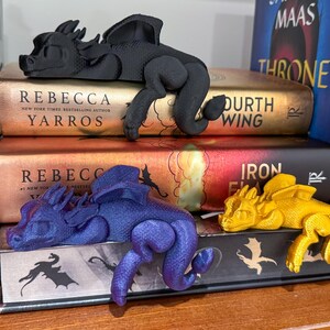 May include: Three dragon bookends in different colors: black, blue, and gold. The bookends are resting on top of a stack of books with titles like "Throne of Glass" and "Iron Flame".