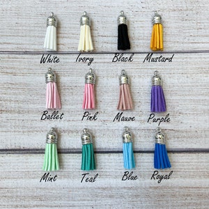 May include: Assortment of 12 tassel charms in various colors, including white, ivory, black, mustard, ballet pink, pink, mauve, purple, mint, teal, blue, and royal blue. Each tassel charm has a silver metal top.