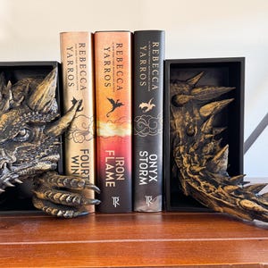 May include: A pair of dragon bookends made of gold-painted resin. The bookends are holding up three fantasy novels by Rebecca Yarros. The books are titled "Onyx Storm", "Iron Flame", and "Fourth Win".
