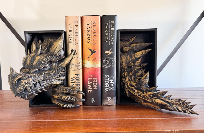 May include: A pair of dragon bookends made of gold-painted resin. The bookends are holding up three fantasy novels by Rebecca Yarros. The books are titled "Onyx Storm", "Iron Flame", and "Fourth Win".
