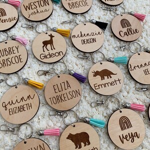 May include: A collection of personalized wooden keychains with various designs and names, including a deer, a dinosaur, a rainbow, and a bear. Each keychain features a small tassel in a variety of colors.