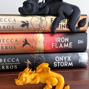 May include: Two dragon bookends, one black and one gold, are resting on a stack of four books. The books are all by the same author, Becca  Rros, and have titles like "Fort Wing", "Iron Flame", and "Onyx Storm".