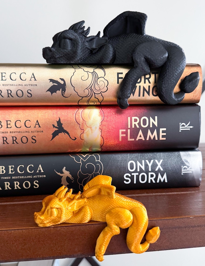 May include: Two dragon bookends, one black and one gold, are resting on a stack of four books. The books are all by the same author, Becca  Rros, and have titles like "Fort Wing", "Iron Flame", and "Onyx Storm".