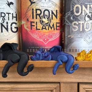 May include: Three 3D printed dragon bookends in black, blue, and gold. The bookends are resting on a wooden shelf in front of three fantasy novels titled "Fourth Wing", "Iron Flame", and "Onyx Storm".